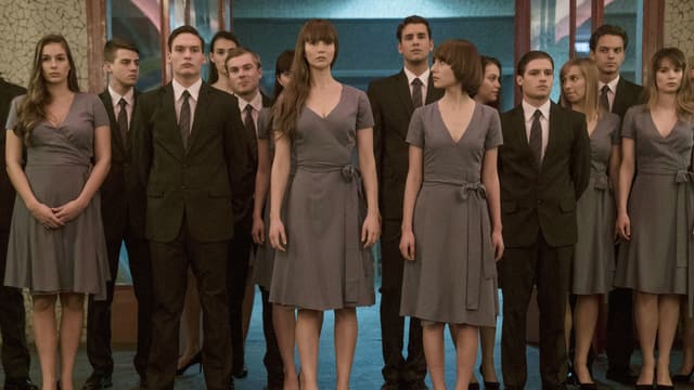 Eine Gruppe junger Menschen stehen im Eingang zu einer Halle. Alle schauen etwas verstört nach vorne oder schauen einander an. Die Männer tragen Anzüge, die Frauen schlichte Kleider. 