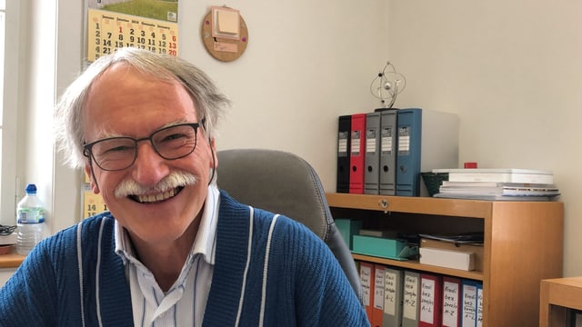 Ein Mann mit grauen Haaren und buschigem Schnauz lächelt in einem Büro in die Kamera. Ein Mann mit grauen Haaren und buschigem Schnauz lächelt in einem Büro in die Kamera.