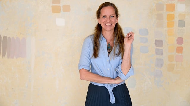 Eine Frau in hellblauem Hemd steht vor einer mit dezenten Farben bemalter, beiger Wand. Eine Frau in hellblauem Hemd steht vor einer mit dezenten Farben bemalter, beiger Wand.