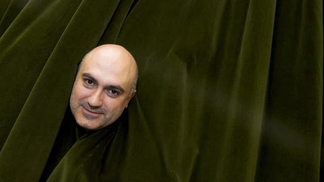 Regisseur Calixto Bieito blickt Betrachter durch Loch in Theatervorhang an. Regisseur Calixto Bieito blickt Betrachter durch Loch in Theatervorhang an.