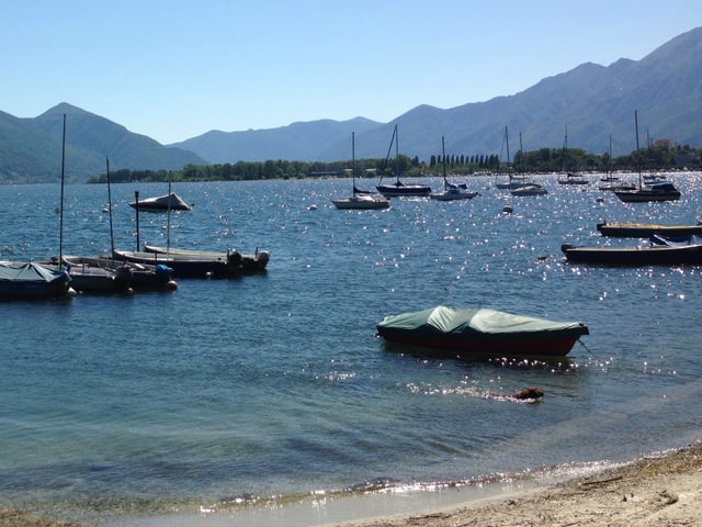 Unten ist knapp noch Sandstrand zu sehen. Im Wasser liegen einige kleine Boote vor Anker. Im Hintergrund liegen die Berge in leichtem Dunst, darüber ist blauer Himmel.