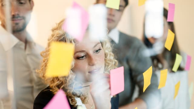 Eine Frau schreibt auf einen Post-It, der mit vielen anderen an einer Glaswand klebt. Eine Frau schreibt auf einen Post-It, der mit vielen anderen an einer Glaswand klebt.