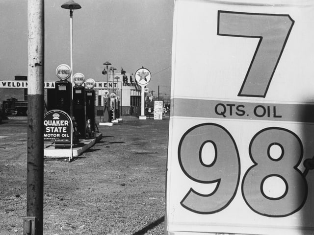 Auf einer schwarz-weissen Fotografie ist eine verlassene Tankstelle zu sehen: Zapfsäulen, rechts ein Schild mit den Ziffern sieben und 98. Auf einer schwarz-weissen Fotografie ist eine verlassene Tankstelle zu sehen: Zapfsäulen, rechts ein Schild mit den Ziffern sieben und 98.