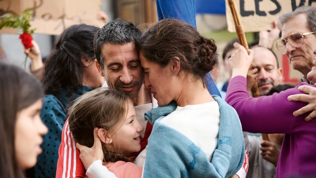 Inmitten einer grösseren Gruppe umarmt sich eine Familie mit Vater, Mutter, Kind. Es scheint eine Wiederzusammenkunft zu sein. Inmitten einer grösseren Gruppe umarmt sich eine Familie mit Vater, Mutter, Kind. Es scheint eine Wiederzusammenkunft zu sein.