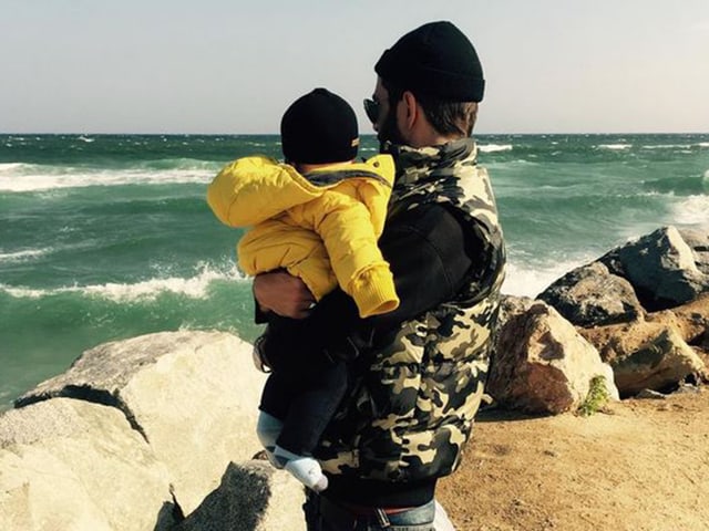 Bligg blickt mit einem Baby in gelber Jacke auf dem Arm aufs Meer. Bligg blickt mit einem Baby in gelber Jacke auf dem Arm aufs Meer.