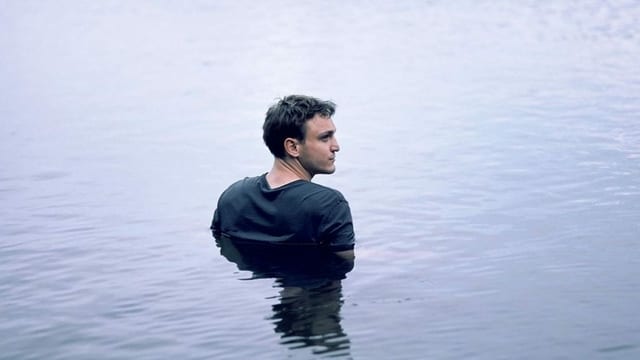 Franz Rogowski steht für eine Filmszene mit Kleidern in einem See. Das Wasser reicht ihm bis zur Brust.