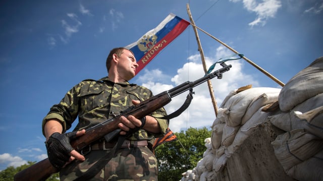 Ein pro-russischer Soldat vor der Russischen Flagge, er hält ein Gewehr in der Hand und blickt in die Ferne. Das Bild wurde in der Froschperspektive aufgenommen. 