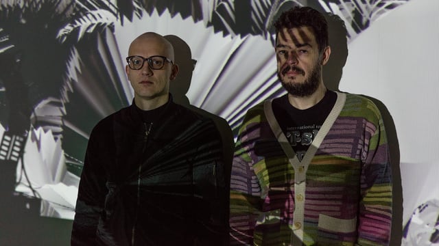 Zwei Männer stehen vor einer Wand, die auf die Schwarzweissbilder von tropischen Pflanzen projeziert werden. Zwei Männer stehen vor einer Wand, die auf die Schwarzweissbilder von tropischen Pflanzen projeziert werden.