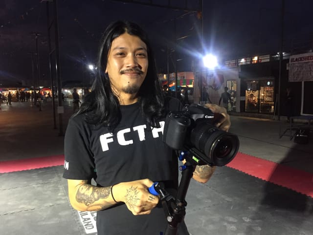 Ein junger Mann mit langen Haaren und einer Profi-Kamera in der Hand. Er trägt ein schwarzes T-Shirt mit dem Logo FCTH (Fight Club Thailand). Ein junger Mann mit langen Haaren und einer Profi-Kamera in der Hand. Er trägt ein schwarzes T-Shirt mit dem Logo FCTH (Fight Club Thailand).