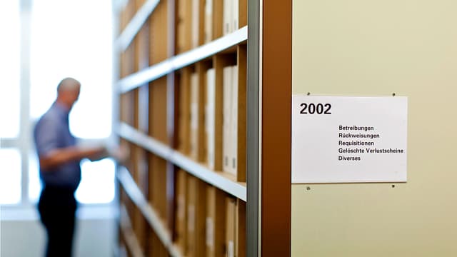 Bild einer Aktenschrank-Wand in einem Betreibungsamt. Im verschwommenen Hintergrund steht ein Betreibungsbeamter am Regal. Bild einer Aktenschrank-Wand in einem Betreibungsamt. Im verschwommenen Hintergrund steht ein Betreibungsbeamter am Regal.