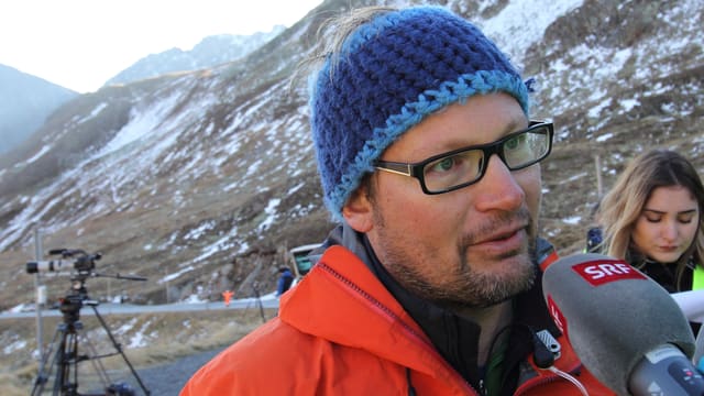 Ein Mann mit einer Strickmütze und orangen Gore-Tex-Jacke steht vor dem Mikrofon. Ein Mann mit einer Strickmütze und orangen Gore-Tex-Jacke steht vor dem Mikrofon.