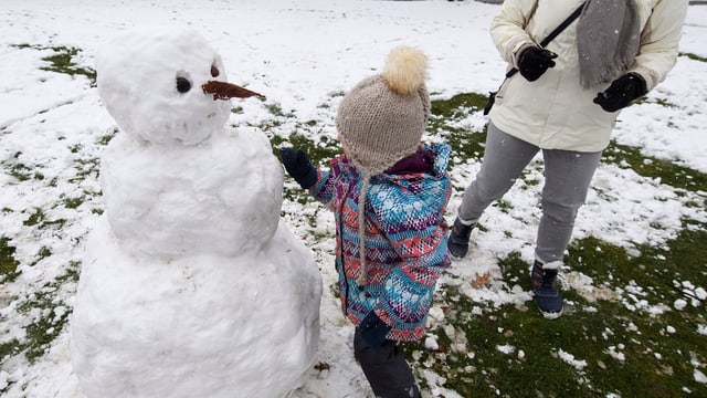 Ein Kind baut einen Schneemann, eine erwachsene Person steht daneben, von der man nur die Beine sieht. Ein Kind baut einen Schneemann, eine erwachsene Person steht daneben, von der man nur die Beine sieht.