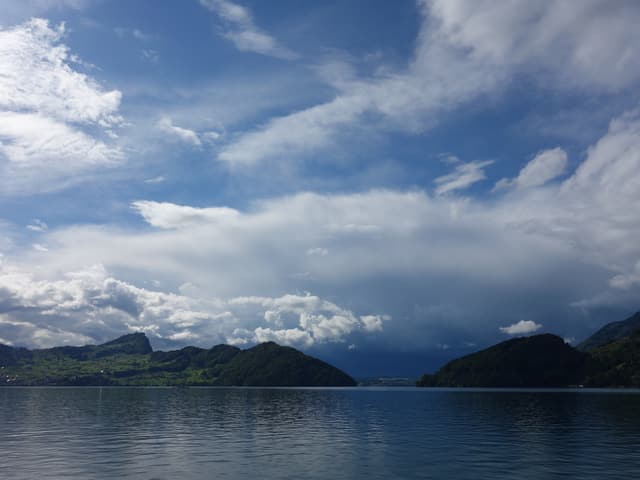 See im Vordergrund, im Hintergrund grüne Berge mit vielen Wolken, aber auch blauem Himmel.