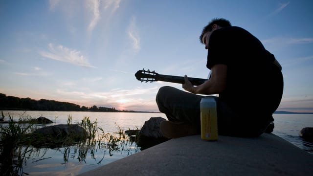 Ein Mann sitzt mit der Gitarre und einem kühlen Getränk am See. Ein Mann sitzt mit der Gitarre und einem kühlen Getränk am See.
