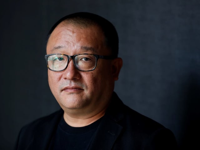 Nahaufnahme: Ein Mann mit asiatischer Herkunft schaut in die Kamera. Er hat kurze, schwarze Haare, trägt eine schwarze Hornbrille, ein schwarzes T-Shirt und darüber ein dunkles, samitges Jakett. Nahaufnahme: Ein Mann mit asiatischer Herkunft schaut in die Kamera. Er hat kurze, schwarze Haare, trägt eine schwarze Hornbrille, ein schwarzes T-Shirt und darüber ein dunkles, samitges Jakett.