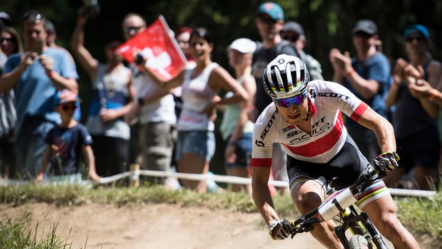 Nino Schurter sin in mountain bike en ina curva. Il terren e natiral e schetg. davostiers ina gruppa d'aspectaturs che applaudeschan. Ina bandiera svizra è da vesair. Nino Schurter sin in mountain bike en ina curva. Il terren e natiral e schetg. davostiers ina gruppa d'aspectaturs che applaudeschan. Ina bandiera svizra è da vesair.