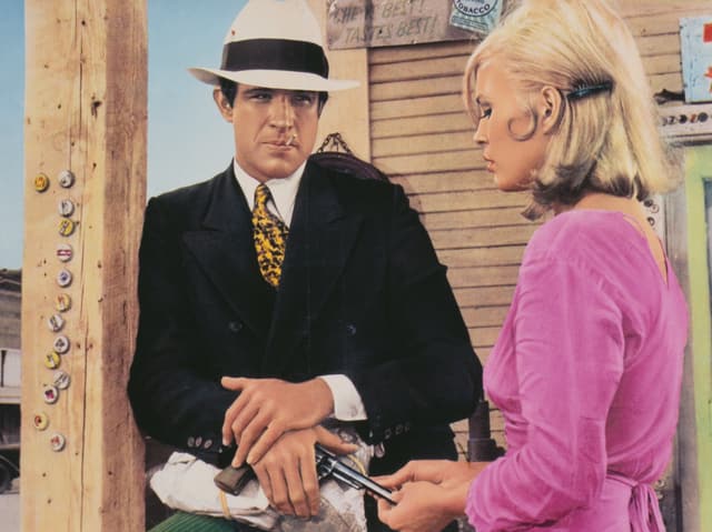 Warren Beatty als Gangster Clyde im Anzug steht neben Gangsterbraut Bonnie, die ein pinkes Kleid trägt.