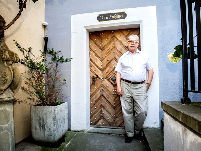 Ein älterer Herr steht vor seiner Haustür. Über der Tür steht "Zum Schützen" angeschrieben. Ein älterer Herr steht vor seiner Haustür. Über der Tür steht "Zum Schützen" angeschrieben.