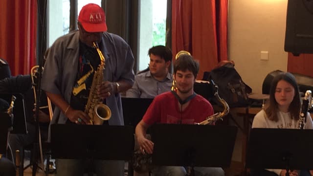 Ein Mann mit Käppchen spielt stehend Posaune, daneben sitzen einige Orchestermitglieder Ein Mann mit Käppchen spielt stehend Posaune, daneben sitzen einige Orchestermitglieder