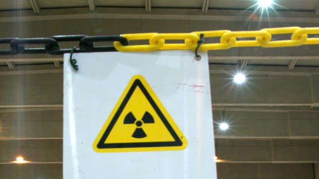 Weisses Warntransparent mit dem Symbol für Radioaktivität in gelbem Dreieck an einer Kette hängend Weisses Warntransparent mit dem Symbol für Radioaktivität in gelbem Dreieck an einer Kette hängend