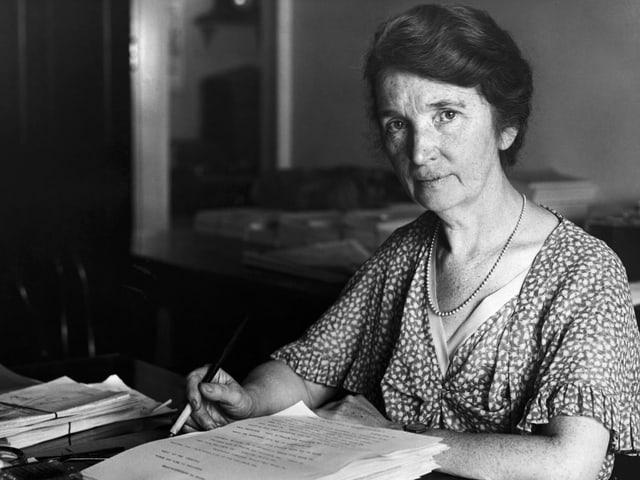 Schwarz-weiss-Bild einer Frau, die mit wachem Blick an einem Schreibtisch sitzt. Schwarz-weiss-Bild einer Frau, die mit wachem Blick an einem Schreibtisch sitzt.