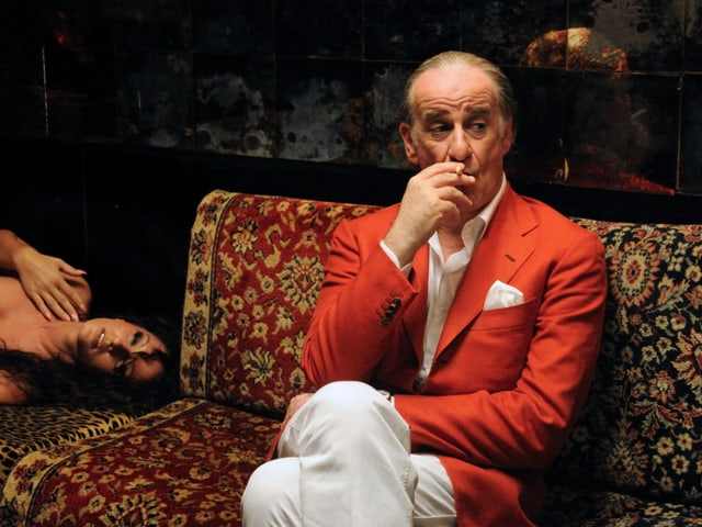 Ein elegant gekleideter Mann sitzt rauchend auf einem Sofa, neben ihm liegt eine nackte Frau. Ein elegant gekleideter Mann sitzt rauchend auf einem Sofa, neben ihm liegt eine nackte Frau.