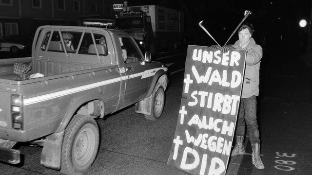 Ein Mann hält ein Plakat, auf dem «Unser Wald stirbt auch wegen dir!» steht, an einer Autostrasse. Ein Mann hält ein Plakat, auf dem «Unser Wald stirbt auch wegen dir!» steht, an einer Autostrasse.