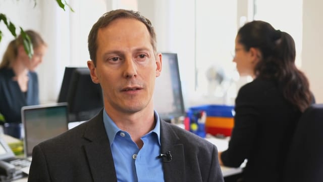 Ein Mann im schwarzen Kittel steht in einem Büro, hinter ihm sitzen zwei Frauen Ein Mann im schwarzen Kittel steht in einem Büro, hinter ihm sitzen zwei Frauen