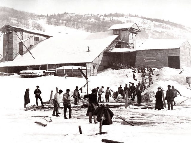 Schwarz-Weiss-Bild: Eine Gruppe Männer und Frauen sticht 1881 Eis, im Hintergrund ein verschneiter Bauernhof.