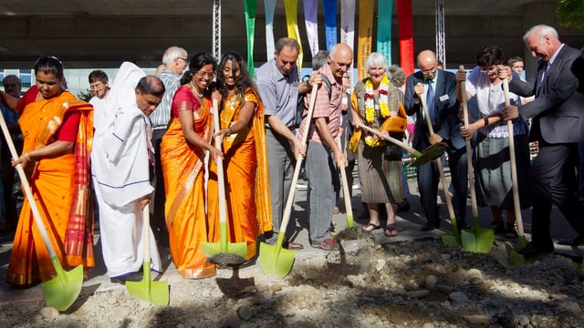 In einer Reihe stehen Frauen in Saris und Männer in geistlichen Kutten. Alle beginnen mit einer Schaufel zu graben. In einer Reihe stehen Frauen in Saris und Männer in geistlichen Kutten. Alle beginnen mit einer Schaufel zu graben.