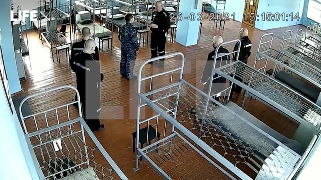 Nawalny mit geschorenem Kopf steht in einem Schlafsaal – Betten ohne Matratzen – vor fünf Wärtern in Uniform.