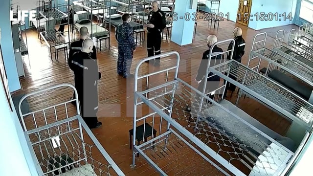 Nawalny mit geschorenem Kopf steht in einem Schlafsaal – Betten ohne Matratzen – vor fünf Wärtern in Uniform. Nawalny mit geschorenem Kopf steht in einem Schlafsaal – Betten ohne Matratzen – vor fünf Wärtern in Uniform.