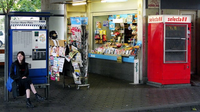 Eine junge Frau sitzt vor einem Schweizer Kiosk. Daneben ist ein roter Selecta-Automat. Eine junge Frau sitzt vor einem Schweizer Kiosk. Daneben ist ein roter Selecta-Automat.
