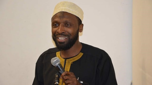 Der Iman hält ein Mikrofon und lächelt. Der Iman hält ein Mikrofon und lächelt.