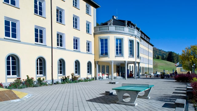 Das Gebäude des Hochalpinen Instituts Ftan. Im Hintergrund Bäume und Wiesen, im Vordergrund ein Tischtennis-Tisch.