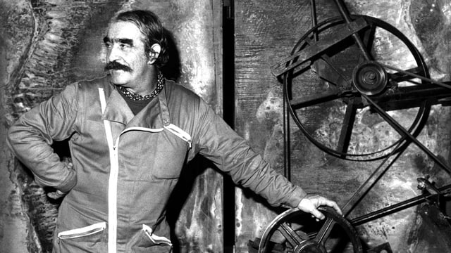 Jean Tinguely steht vor einem Kunstwerk aus Rädern. Jean Tinguely steht vor einem Kunstwerk aus Rädern.