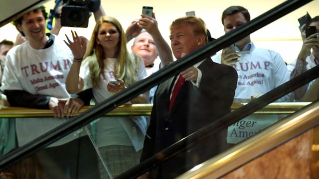 Donald Trump auf einer Rolltreppe. Menschen winken dahinter und eine Kamera filmt ihn. Er hebt den Daumen hoch.