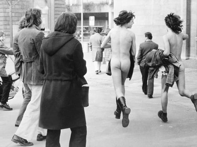 Zwei nackte Frauen rennen durch eine Strasse, Passanten lachen ihnen zu. Zwei nackte Frauen rennen durch eine Strasse, Passanten lachen ihnen zu.