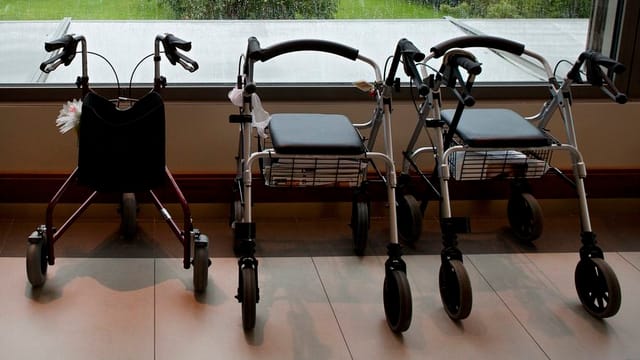 Drei Rollatoren stehen in einem Pflegeheim auf einem Parkettboden vor einem Fenster. Drei Rollatoren stehen in einem Pflegeheim auf einem Parkettboden vor einem Fenster.