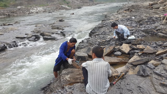 Drei Männer sitzen an einem Fluss und bearbeiten Steinplatten mit Hämmern Drei Männer sitzen an einem Fluss und bearbeiten Steinplatten mit Hämmern