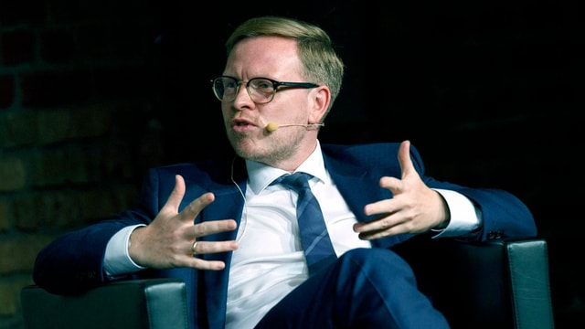 Ein Mann in Anzug sitzt gestikulierend auf einem schwarzen Ledersessel. Ein Mann in Anzug sitzt gestikulierend auf einem schwarzen Ledersessel.
