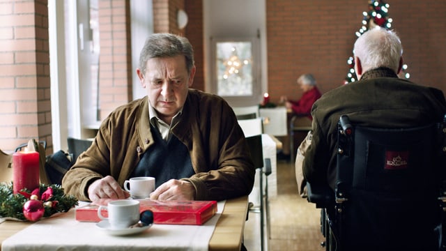Ein einsamer Mann an einem Tisch im Altersheim, ein anderer fährt im Rollstuhl an ihm vorbei Ein einsamer Mann an einem Tisch im Altersheim, ein anderer fährt im Rollstuhl an ihm vorbei