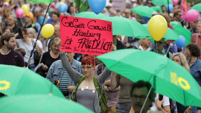 Frau inmitten einer Demostration mit Schil in der Hand, das die Aufschift trägt: «Gegen Gewalt, Hamburg ist friedelich» Frau inmitten einer Demostration mit Schil in der Hand, das die Aufschift trägt: «Gegen Gewalt, Hamburg ist friedelich»