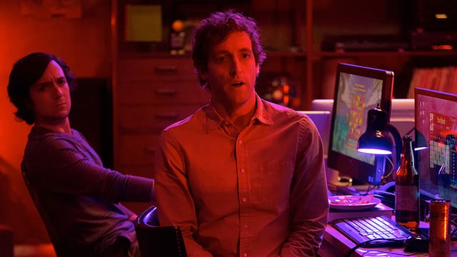 Ein in Rotlicht getauchter junger Mann schaut überrascht in die Kamera, hinter ihm ein am Computer sitzender zweiter junger Mann, dessen Blick eher skeptisch ist.