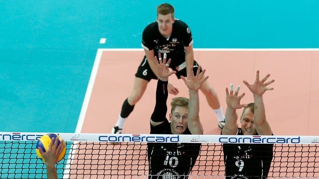 2 Männer springen hoch und heben die Hände, während eine Hand versucht den Ball über das Netz zu spielen. 2 Männer springen hoch und heben die Hände, während eine Hand versucht den Ball über das Netz zu spielen.