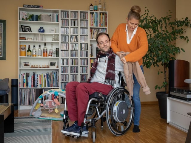Eine stehende Frau zieht einem Mann im Rollstuhl die Jacke an. Eine stehende Frau zieht einem Mann im Rollstuhl die Jacke an.