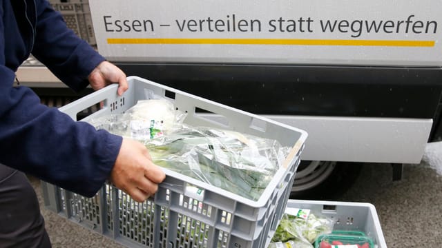Person hält eine Kiste mit Gemüse, im Hintergrund der Schriftzug "Essen verteilen statt wegwerfen" Person hält eine Kiste mit Gemüse, im Hintergrund der Schriftzug "Essen verteilen statt wegwerfen"