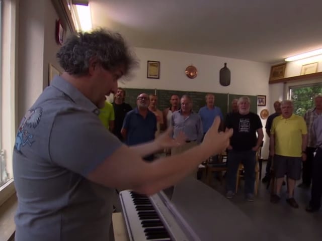 Ein Dirigent an einem Keyboard vor einer Gruppe singender Männer Ein Dirigent an einem Keyboard vor einer Gruppe singender Männer