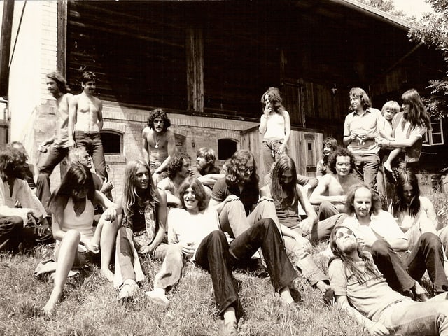 Eine grössere Gruppe von Menschen mit langen Haaren auf einer Wiese vor einem Holzhaus. Eine grössere Gruppe von Menschen mit langen Haaren auf einer Wiese vor einem Holzhaus.