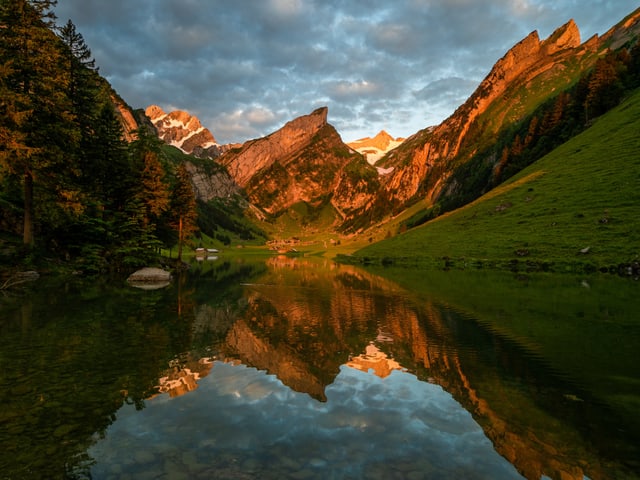 Blick über einen Bergsee. Die Berge dahinter leuchten im Morgenrot und spiegeln sich im Wasser.